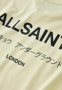 Krémová látka s čiastočne viditeľným čiernym textom "ALLSAINT", japonské znaky pod ním a pod nimi vytlačený nápis "LONDON".