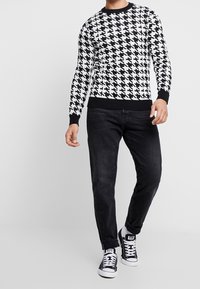 Svetr s černobílým houndstooth vzorem, kulatým výstřihem a žebrovanými manžetami a lemem, v kombinaci s tmavými džíny a teniskami.