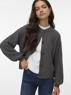 Cardigan gris en maille avec manches longues bouffantes, doté d'un col rond et de cinq gros boutons sur le devant. Porté sur un haut blanc.