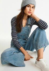 Ljust blå denimsoverall med vida ben, i kombination med en svart- och vitrandig långärmad tröja och vita sneakers. Grå stickad beanie.