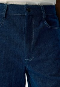 Primo piano della vita di un paio di jeans in denim blu scuro con bottone, passanti per cintura e cuciture visibili sopra l'area dell'anca e della tasca.