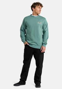 Sweatshirt vert menthe avec un col rond, des poignets côtelés et une ceinture côtelée. Présente un petit logo imprimé sur la poitrine. Porté avec un pantalon noir et des chaussures.
