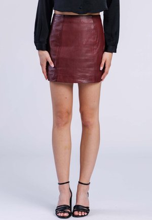 Vrouw draagt een donkerrode leren minirok, zwarte top met lange mouwen en zwarte hoge sandalen met bandjes, staand tegen een effen achtergrond.