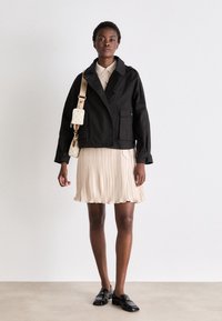 Veste noire courte avec poches avant, portée avec une robe plissée beige et des chaussures noires. Les accessoires comprennent un petit sac crème avec une bandoulière.