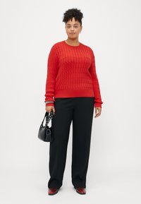 Rød kabelstrikket genser med ribbede mansjetter, parret med svarte vide bukser. Tilbehøret inkluderer en svart håndveske med naglede stropper.