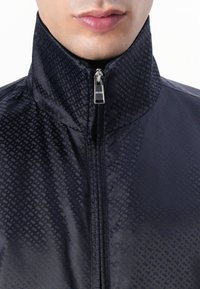 Jūrinės spalvos zip-up striukė su aukštu apykakle, turinti geometrinį raštą. Pagaminta iš lygaus, blizgaus audinio su metaliniu užtrauktuku.