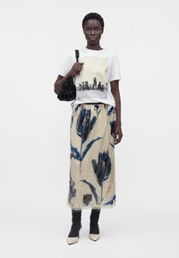 Femme portant un t-shirt blanc avec des images de chiens, une jupe beige avec de grands motifs floraux bleus, des chaussettes noires, des talons blancs et tenant un sac à main noir.