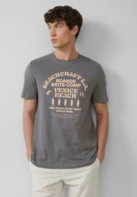 Grijze t-shirt met korte mouwen heeft een groot bedrukt ontwerp in perzik, met de tekst "Beachcraft L.A. Venice Beach" en afbeeldingen van surfplanken.
