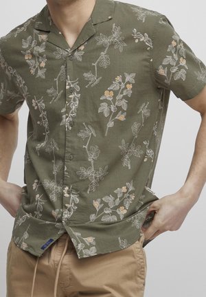 Man draagt een olijfgroen overhemd met korte mouwen en een witte en oranje bloemenprint, gecombineerd met beige broek, handen in de zakken.