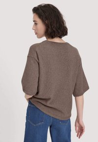 Maglione marrone lavorato a maglia con maniche corte, vestibilità rilassata, colletto tondo e orlo a coste, indossato sopra jeans blu. La texture sembra morbida.