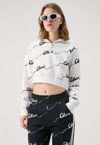 Sudadera recortada blanca con patrón de texto negro en forma de ondas y cremallera al frente, combinada con pantalones jogger negros que presentan el mismo patrón de texto. Gafas de sol blancas.