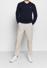 Námornícky svetr s véčkovým výstřihem v kombinaci s béžovými kalhotami a bílými teniskami. Svetr má na hrudi malé zelené logo; hladká textura po celém povrchu.