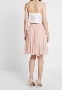 Nainen, joka on pukeutunut valkoiseen hihattomaan puseroon, vaaleanpunaiseen laskostettuun midi-hameeseen ja vaaleanpunaisiin korkokenkiin, seisoo selkä kameraan päin.