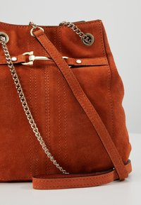 Sac seau en daim orange avec une bandoulière chaîne et cuir. Comprend des ferrures dorées, des coutures latérales et une fermeture à cordon coulissant.