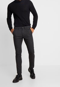 Pantalones gris de cuadros con un corte entallado, combinados con una blusa negra de mangas largas. Unos zapatos negros de cordones completan el conjunto.