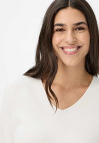 Weißes Strickoberteil mit V-Ausschnitt. Der Stoff hat ein strukturiertes Muster, während das Model lange, dunkle Haare und einen lächelnden Gesichtsausdruck hat.