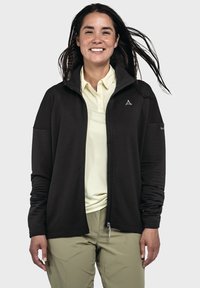 Schwarze Zip-Jacke mit gerippter Textur, verfügt über einen hohen Kragen und ein Logo auf der Brust, kombiniert mit einem hellgelben Hemd darunter.