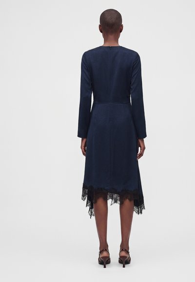 Personne aux cheveux courts portant une robe bleu foncé à manches longues avec un bord en dentelle à l'ourlet et des chaussures à talons hauts foncées, debout de dos.