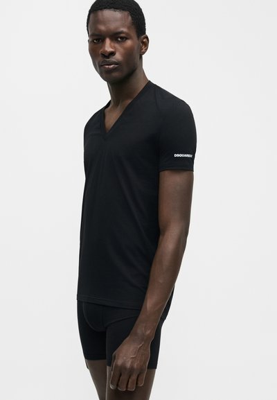 Άνδρας που φοράει μαύρο V-neck T-shirt με το κείμενο «DSQUARED2» στο μανίκι και μαύρα εφαρμοστά σορτς, στέκεται μπροστά σε απλό φωτεινό φόντο.