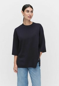 Marineblauwe oversized trui met een ronde hals, korte mouwen en zijsplitten, vervaardigd uit een zachte brei-stof. Gecombineerd met lichtblauwe jeans.