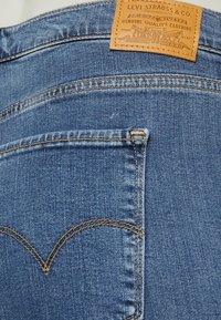 Vaqueros de denim azul con un acabado texturizado, con costuras naranjas, bolsillos traseros y un parche de cuero marrón con detalles de la marca.