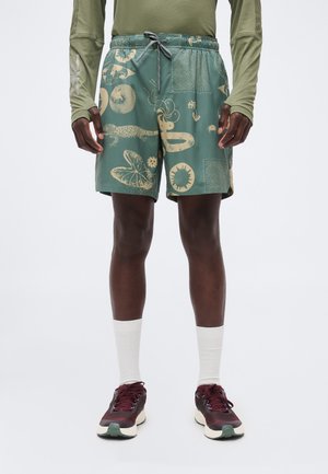 Mann iført grønne shorts med snor og beige naturmotivtrykk, langermet olivengrønn skjorte, hvite sokker og burgunder joggesko, står.