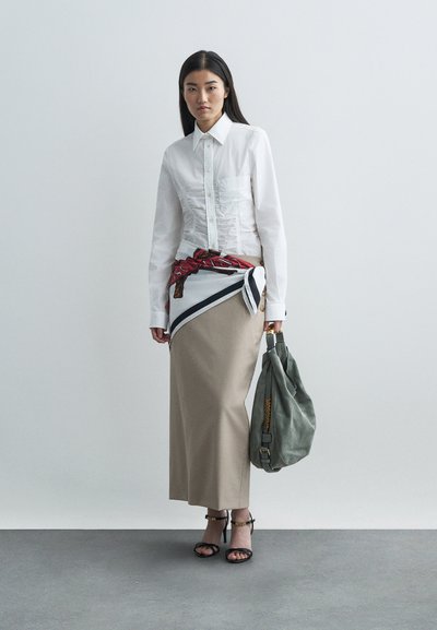 Žena v bielej riasenej blúzke, béžovej dlhéj sukni s viazanou vzorovanou šatkou, čiernych sandáloch na podpätku, ktorá drží veľkú sivú kabelku.