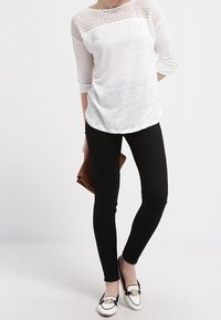 Bílý, volný top s krajkovým panelem na ramenech, doplněný černými skinny džíny a černými mokasíny s dekorativním zlatým akcentem.