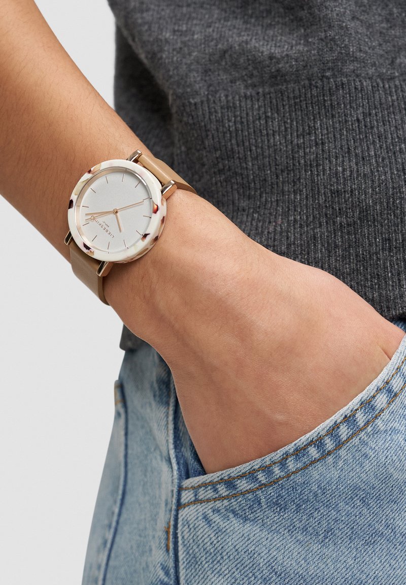 Runde silberne Uhr mit beigem Lederarmband und mehrfarbigem Schildpatt-Lünettenring, zeigt ein minimalistisches Zifferblatt mit metallischen Stundenmarkierungen.