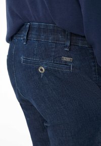 Ansicht von hinten auf eine Person, die dunkelblaue Jeans mit einer geknöpften Gesäßtasche und einem kleinen Metalletikett mit der Aufschrift "Eurex" trägt.