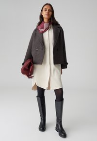 Cappotto doppiopetto in misto lana grigio scuro su un vestito crema. Accessoriato con una sciarpa bordeaux con righe bianche. Stivali neri fino al ginocchio.