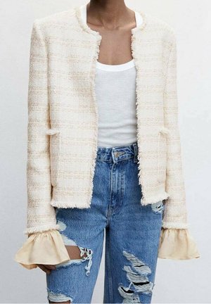 Person iført en hvid tekstureret jakke over en hvid tanktop med flagrende beige manchetter og revnede blå jeans.