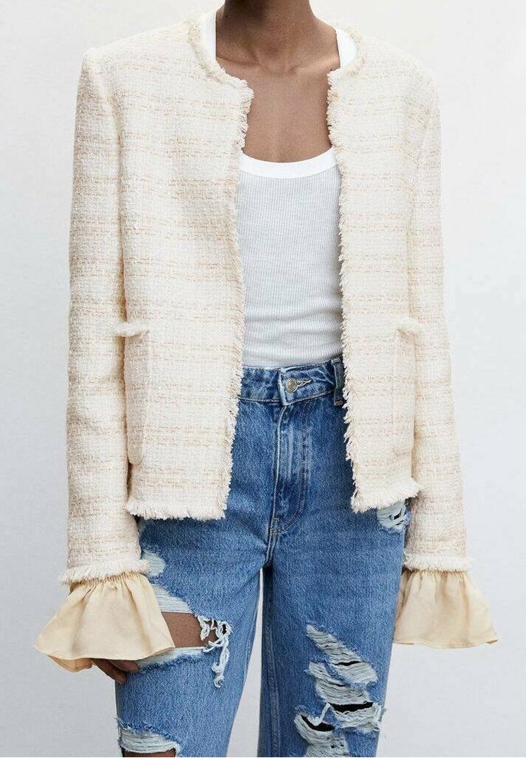 Personne portant une veste blanche texturée sur un débardeur blanc avec des poignets beige évasés et un jean bleu déchiré.