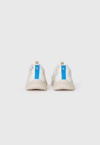 Zapatillas deportivas blancas con suelas beige, que cuentan con un acento azul en el talón y un pequeño lazo para ponérselas fácilmente. Diseño de parte superior texturizada.