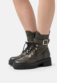 Botas de tobillo de cuero verde oliva con una suela negra gruesa, que cuentan con ojales en tono dorado, cordones negros y un diseño de puño con hebilla.