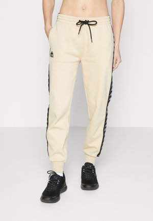Personne portant un pantalon de jogging beige avec des bandes noires sur les côtés et un logo, associé à des baskets noires, debout sur un fond clair uni.