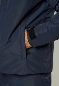 Hand in der Tasche einer navyblauen Jacke mit verstellbarem Velcro-Manschette, gezeigt von der Taille bis zur Mitte des Oberschenkels.