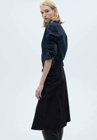 Mörk denimskjorta med uppvikta ärmar, som kombineras med en lång, svart denimkjol. Modellen poserar i profil och visar plaggets form och textur.