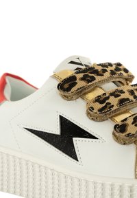 Baskets en cuir blanc avec un éclair en cuir noir, des sangles Velcro dorées et imprimées léopard, un accent rouge au talon et une semelle en caoutchouc texturé.