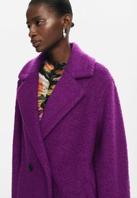 Abrigo texturizado en púrpura con cuello de muesca, diseño de doble botonadura y botones negros; llevado sobre una blusa de patrón colorido.