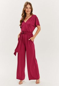 Fuchsiový overal s širokými nohavicemi, krátkými rukávy a výstřihem do V. Má opasek v pase a boční kapsy, vyrobený z hladké, splývavé tkaniny.