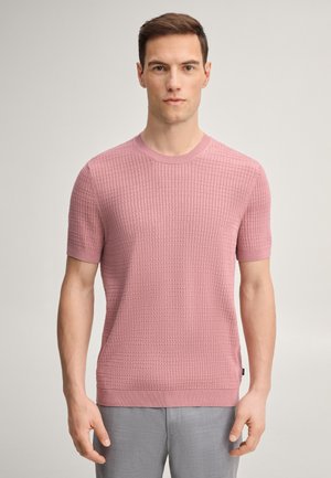 Ein Mann steht frontal, trägt einen strukturierten rosa Kurzarm-Pullover und hellgraue Hosen vor einem schlichten, hellen Hintergrund.
