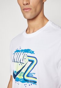 Valge puuvillane T-särk, millel on sinine ja roheline graafiline print, kus on kirjas sõna "NIKE" ja abstraktsed tilgakujulised mustrid. Ümmargune kaelus, lühikesed varrukad.