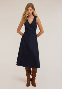 Denim jurk in donkerblauw met een V-hals, mouwloos ontwerp en geplooide rok. G gedragen met lange glanzende bruine laarzen. Soepele textuur en een perfecte pasvorm.