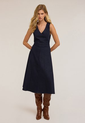 Denim jurk in donkerblauw met een V-hals, mouwloos ontwerp en geplooide rok. G gedragen met lange glanzende bruine laarzen. Soepele textuur en een perfecte pasvorm.