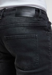 Artiškinis juodų džinsų kišenės ir juosmens kraštas su juoda odine etiketė "ZY Premium", dėvimi su baltu marškinėliu.