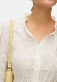 Vit texturerad blus med volangkrage och knappstängning. En mjuk gul axelrem är synlig över axeln.