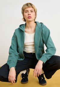 Grønn zip-up hettegenser med en myk tekstur, båret over en hvit topp. Kombinert med svarte bukser og svarte joggesko med brun såle.