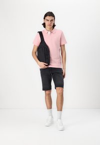 Sac à bandoulière noir porté sur l'épaule gauche avec un polo rose clair, assorti à un short sombre et des baskets blanches. Fond uni.
