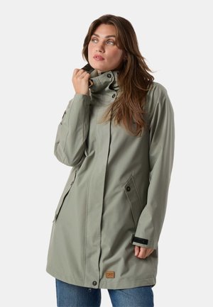 Femme ajustant le col d'un long manteau imperméable vert clair avec poches boutonnées, debout devant un fond uni.